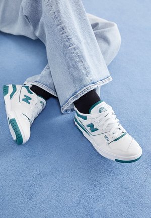 Hvide sneakers med grønne accenter, der har en tekstureret overdel og en gummisål. Kombineret med lyseblå denim, på en blå baggrund.