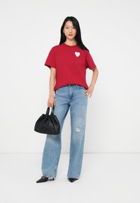 Femeie în picioare, îmbrăcată cu un tricou roșu cu un logo alb în formă de inimă, blugi deschiși la culoare cu găuri, pantofi negri cu vârful ascuțit și ținând o poșetă neagră.