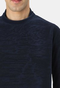 Maglione blu navy con scollo tondo, caratterizzato da una superficie strutturata e sottili motivi a onde. Realizzato in tessuto morbido ed elastico.