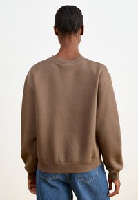 Brown, oversized sweatshirt with a crew neckline and ribbed cuffs. Smooth texture, casual fit. Worn with blue denim jeans.