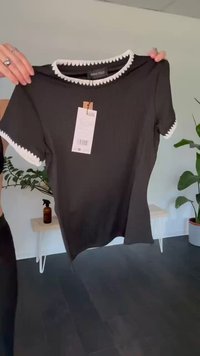 Black short-sleeve ribbed top with white scalloped trim on collar and sleeves, held by hands with a price tag attached.