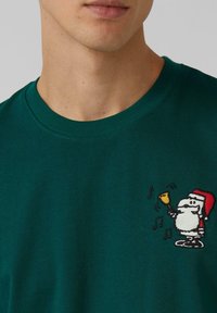 Man wearing dark green shirt with small embroidered cartoon Santa ringing a bell and musical notes on chest.