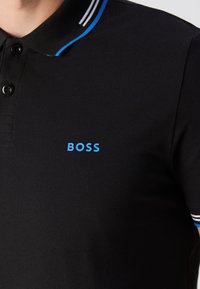 Polo noir avec col rayé bleu et blanc et petit logo bleu « BOSS » sur la poitrine.