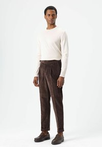 Chemise à manches longues beige clair, pantalon en velours côtelé marron avec plis à l'avant, et mocassins marron foncé, sur un fond blanc uni.