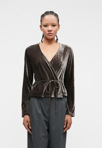 Top din catifea de culoare maro închis, cu mâneci lungi, decolteu în V, curea la talie legată de sine și o textură moale și fină. Asortat cu pantaloni gri croiți.