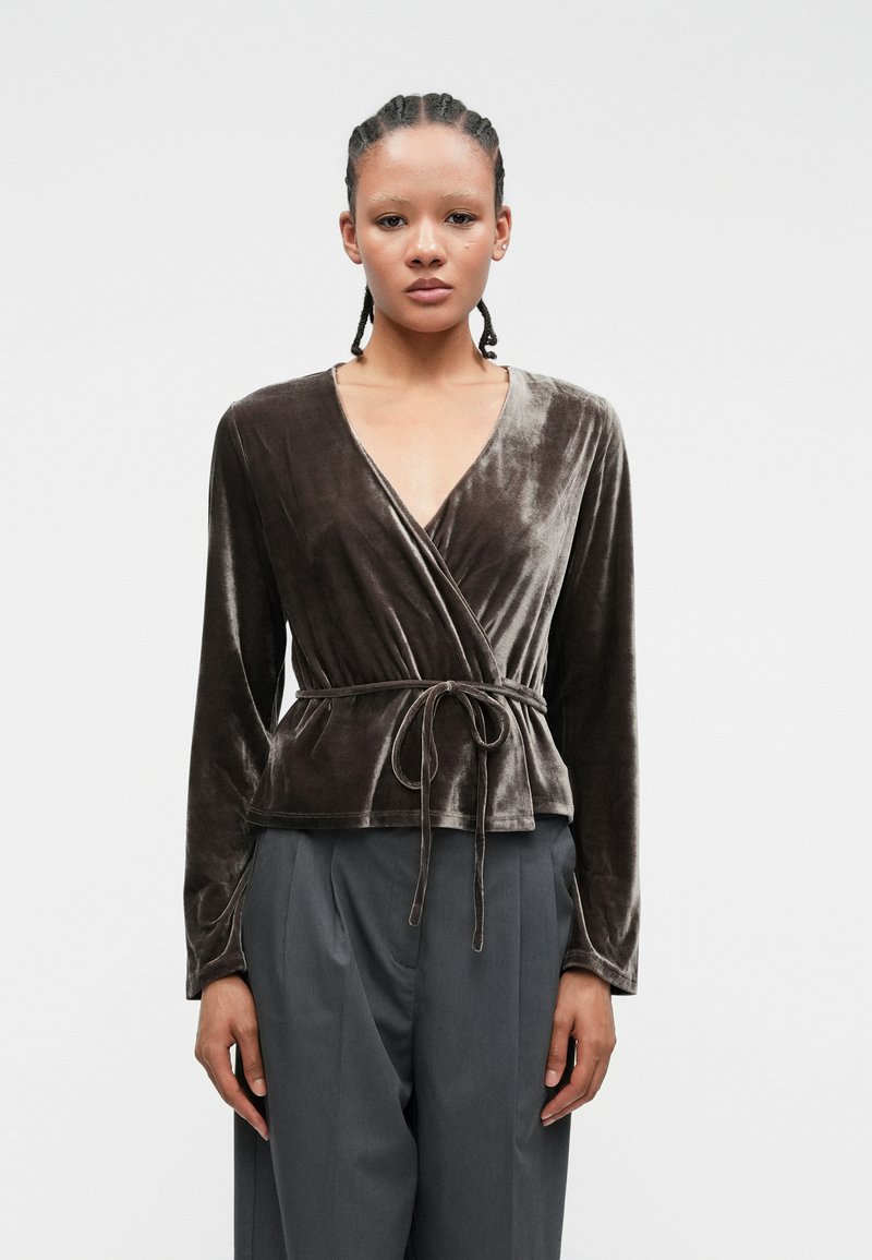 Top din catifea de culoare maro închis, cu mâneci lungi, decolteu în V, curea la talie legată de sine și o textură moale și fină. Asortat cu pantaloni gri croiți.
