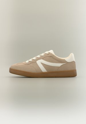Beige sneakers med hvide snørebånd, hvid sidekant, hvid hælblik og brun gummisål på neutral baggrund.