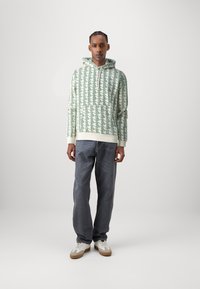 Felpa con cappuccio verde e crema con un motivo geometrico, abbinata a jeans grigio scuro e sneakers bianche. Tessuto in cotone, design casual.