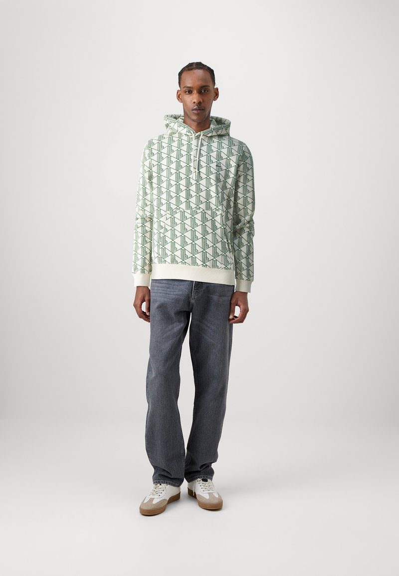 Sudadera con capucha en verde y crema con un patrón geométrico, combinada con jeans gris oscuro y zapatillas blancas. Material de algodón, diseño casual.
