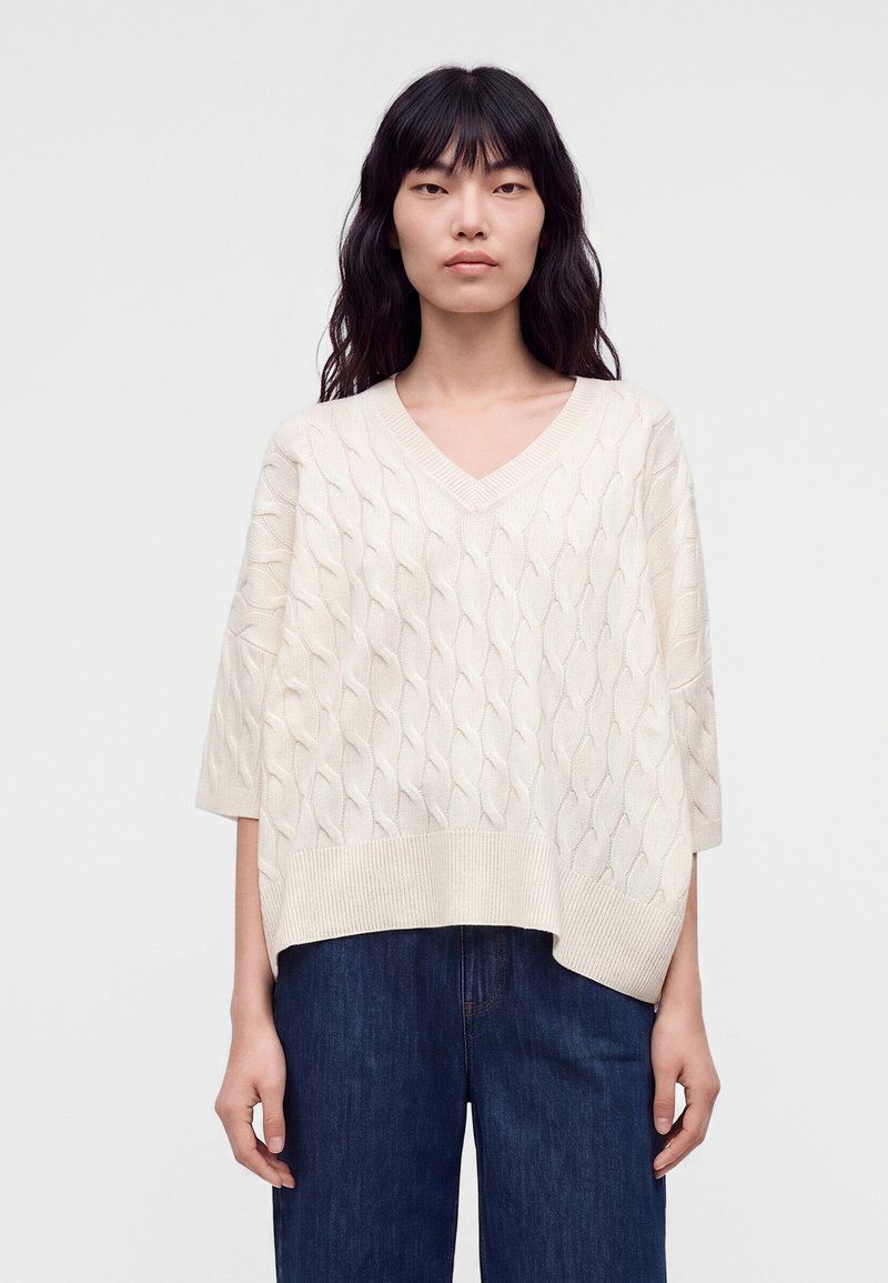 Femme aux longs cheveux noirs portant un pull ample blanc à col en V en maille torsadée et un jean bleu foncé, debout devant un fond clair uni.