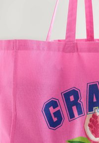 Borsa tote in tela rosa con manici lunghi, caratterizzata da lettere blu in grassetto che formano la parola "GRAT", e un design stampato di un melograno e foglie nella parte inferiore.