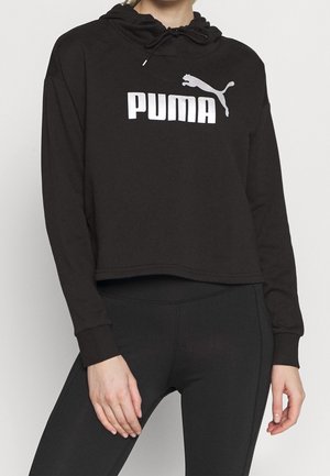 Osoba ubrana w czarną krótka bluzę z kapturem z białym logo Puma oraz czarne dopasowane legginsy, stojąca na jednolitym jasnym tle.