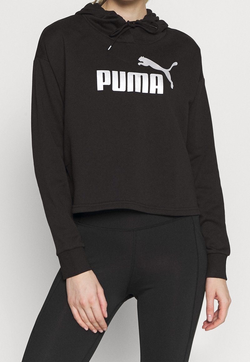 Personne portant un sweat à capuche court noir avec logo Puma blanc et des leggings noirs moulants, debout devant un fond clair uni.