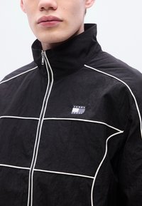 Μαύρο μπουφάν με ψηλό γιακά, λευκές λεπτομέρειες και μικρό λογότυπο "TOMMY JEANS". Διαθέτει φερμουάρ και υφή υφάσματος.