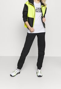 Sportovní oblečení zahrnující černou a žlutou bundu, bílý tričko s logem, černé kalhoty a bílé tenisky se žlutými akcenty.