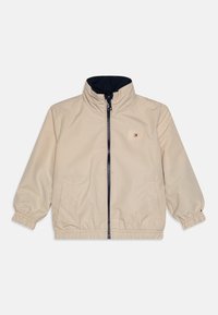 Veste beige à fermeture éclair avec poignets et ourlet élastiques, col montant doublé d'un tissu sombre, petite étiquette logo rectangulaire sur la poitrine.