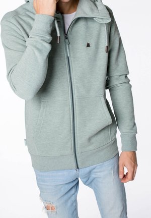 Hombre con sudadera con capucha verde claro con cremallera y bolsillos delanteros, y jeans azules claros rotos, con la mano cerca de la cara.