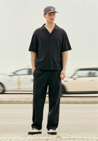 Un jeune homme se tient sur le trottoir, portant une chemise noire à manches courtes, un pantalon noir, une casquette grise et des chaussures blanches, avec des voitures qui passent en arrière-plan.
