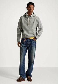 Νεαρός άνδρας φοράει γκρι φούτερ, κίτρινο πουκάμισο, ξεθωριασμένα μπλε τζιν, καφέ παπούτσια, στέκεται με το ένα χέρι στην τσέπη σε λευκό φόντο.