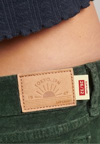 Pantalons pattes d'eph verts avec une surface texturée. Étiquette en cuir marron affichant "Tokyo, JPN" et un graphique de soleil, taille plus 26/32.