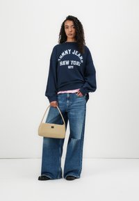 Felpa navy con logo "TOMMY JEANS NEW YORK CITY", abbinata a una maglietta sottile rosa chiaro e jeans a gamba larga in denim. Borsa beige, scarpe nere.