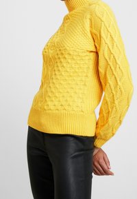 Pull en maille jaune avec col montant, motifs texturés et poignets côtelés, assorti à un pantalon noir pour un contraste saisissant.