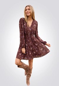 Braunes Blumenkleid mit langen Ärmeln und V-Ausschnitt, tailliert in der Taille, kombiniert mit beigen Kniehochstiefeln. Leichter, fließender Stoff.