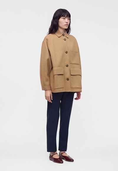 Γυναίκα με καμηλό καφέ oversized μπουφάν με μεγάλες τσεπές με κουμπιά, ναυτικό μπλε ίσια παντελόνια και μπορντό παπούτσια Mary Jane, στέκεται μπροστά σε λευκό φόντο.