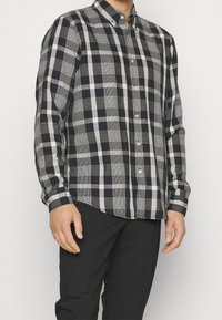 Koszula w kratę z długim rękawem w kolorach czarnym, szarym i białym. Posiada kołnierzyk na guziki, guziki z przodu oraz zaokrąglony dół. Wykonana z bawełnianej tkaniny.