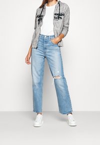 Kvinde i lyseblå ripped jeans, hvide sneakers, hvid t-shirt og grå hættetrøje med sort tekst, stående med den ene hånd i lommen.