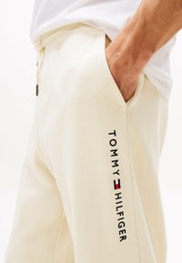 Pantalones de chándal beige de tejido suave con cintura elástica y cordón. Presenta un texto de logo negro "TOMMY HILFIGER" en el lateral.