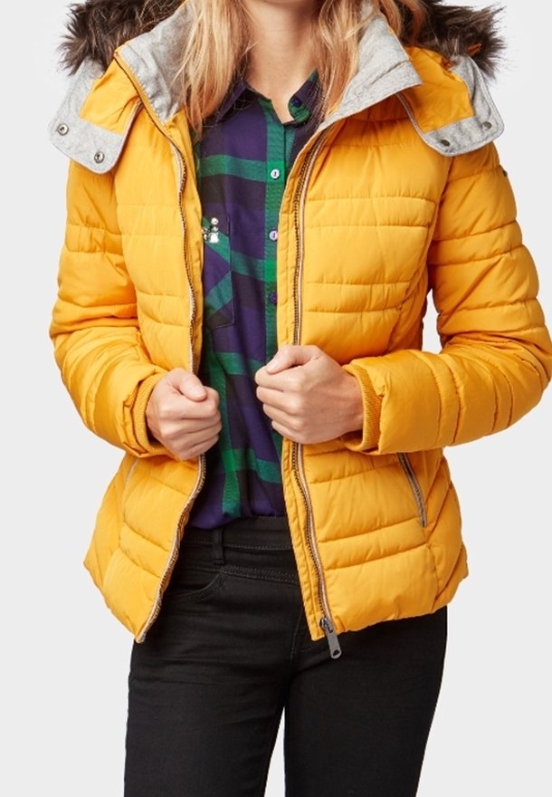 Personne portant une veste matelassée jaune vif par-dessus une chemise à carreaux bleue et verte, tenant la veste ouverte à l'avant, pantalon noir visible.