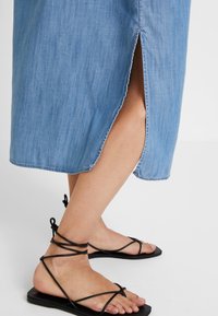 Denim midi-klänning i ljusblå med sidoutskärning. Svarta sandaler med remmar och platt sula. Slät tyg med subtil textur.