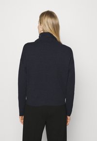 Tummansininen pooloneule, jossa on pitkät hihat, rento istuvuus, pehmeä rakenne ja hieman lyhennetty helma. Yhdistetty mustiin housuihin.