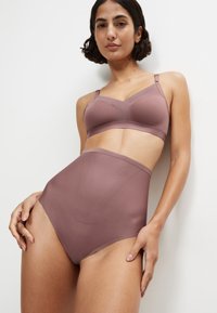 Vrouw die mauve hooggetailleerde corrigerende slips en bijpassende beha draagt, staand tegen een witte muur, met haar handen op haar heupen.