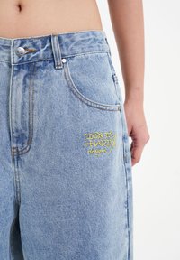 Los vaqueros de denim azul claro presentan una cintura alta, un diseño clásico de cinco bolsillos y texto bordado en amarillo en el muslo izquierdo.