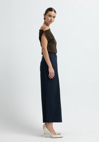 Top marrone senza spalle, pantaloni larghi blu navy e scarpe con tacco alto color crema. Design minimalista con texture lisce e linee pulite.