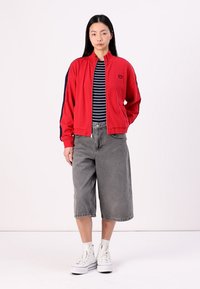 Giacca rossa con accenti blu navy, dotata di chiusura con zip e logo. Camicia a righe sotto, pantaloni corti grigi larghi e scarpe da ginnastica bianche.