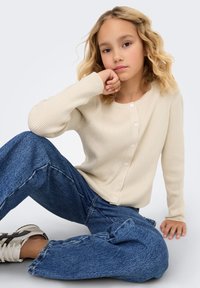 Krämfärgad ribbad kofta med rund halsringning och knappstängning, kombinerad med löst sittande blåa denimjeans och vita sneakers.