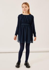 Abito navale con motivo a pois, vita elastica e bordi in pizzo. Abbinato a collant coordinati navy e scarpe in vernice nera.