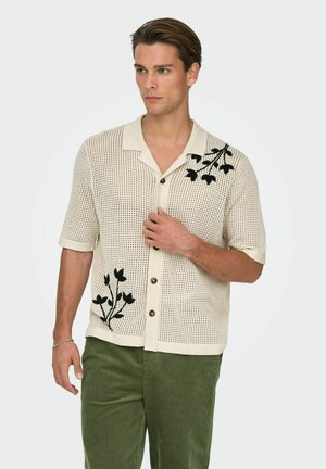 Jeune homme portant une chemise en tricot beige à manches courtes avec une broderie florale noire et un pantalon en velours côtelé vert olive, regardant sur le côté.