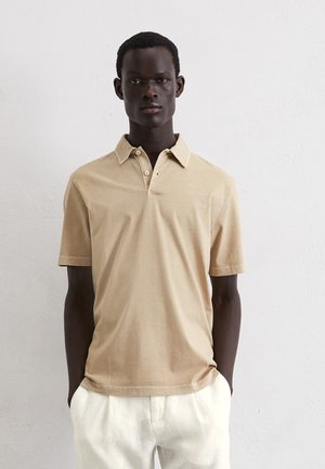 Tan polo shirt made of soft fabric, featuring a collar and three-button placket, paired with light-colored trousers, against a plain background.
