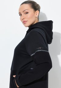 Sudadera negra con textura acanalada, que cuenta con un bolsillo frontal y un acento reflectante en la manga. Logo estiloso en el brazo.