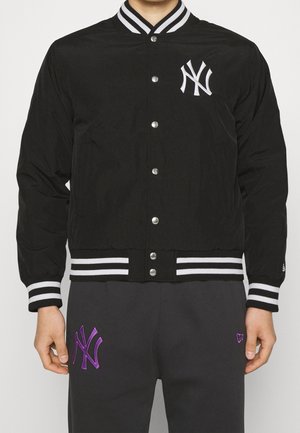 Persoon die een zwarte varsityjas met knopen draagt, met een wit gestreepte kraag en manchetten, en zwarte broek met paarse NY-teamlogo's.