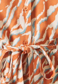 Tkanina s oranžovým a bílým vzorem a modrými akcenty. Obsahuje zapínání na knoflíky, oranžový knoflík a detail s uvázaným pasem.