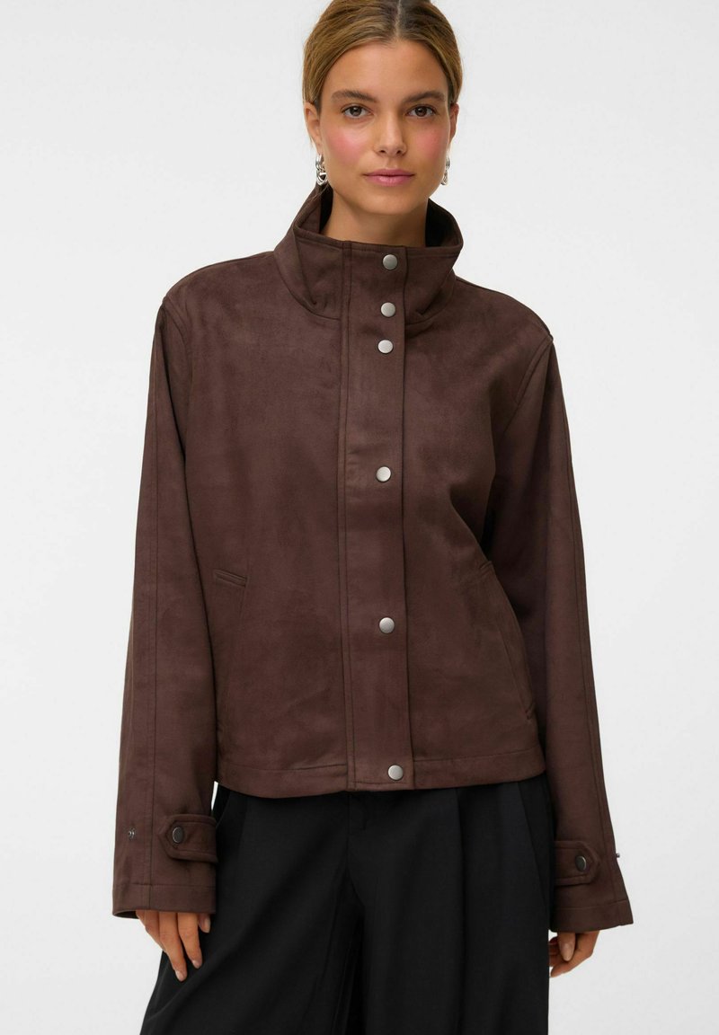 Femme portant une veste marron boutonnée à col montant avec des manches longues et un pantalon noir, regardant droit devant avec une expression neutre.