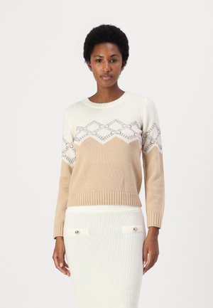 Maglione lavorato a maglia con blocchi di colore crema e beige. Presenta un motivo geometrico sul petto, colletto e polsini a coste. Indossato con una gonna bianca dalla trama testurizzata.