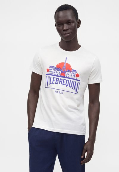 Άνδρας που φοράει λευκό μπλουζάκι με γραφικό της γραμμής ορίζοντα του Παρισιού και του Πύργου του Άιφελ, μπλε κείμενο "Vilebrequin Paris", συνδυασμένο με σκούρο μπλε παντελόνι.