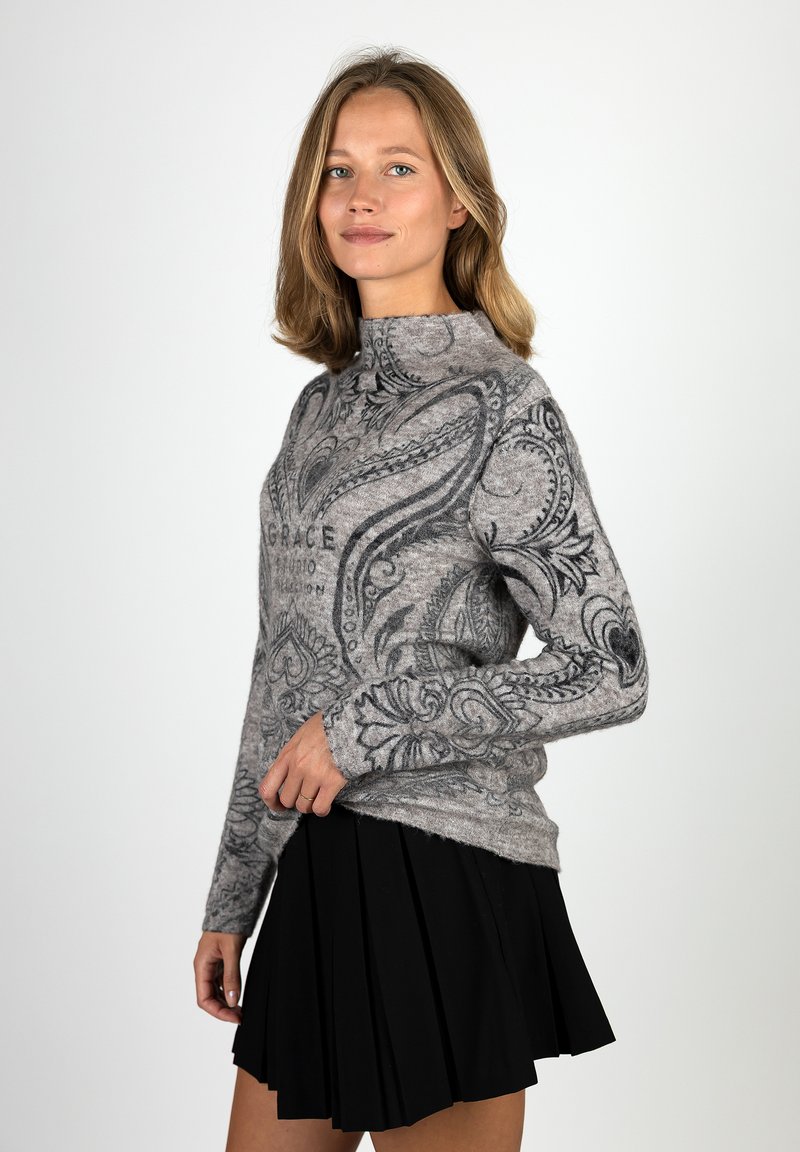 Woman with shoulder-length blonde hair wearing a gray patterned sweater and a black pleated skirt, standing against a plain background.