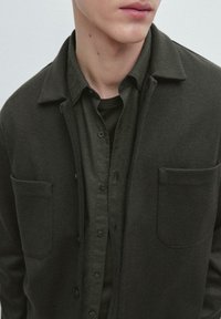 Uomo che indossa una camicia a strati verde scuro, camicia abbottonata e giacca con grandi tasche sul petto, tagliata al collo e in vita.